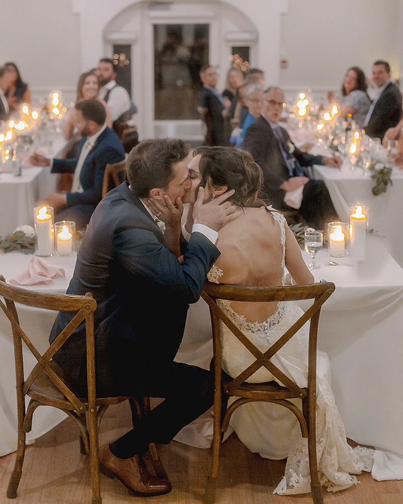 wedding couple is kissing at the reception table surrounded by their families , candlelit dinner