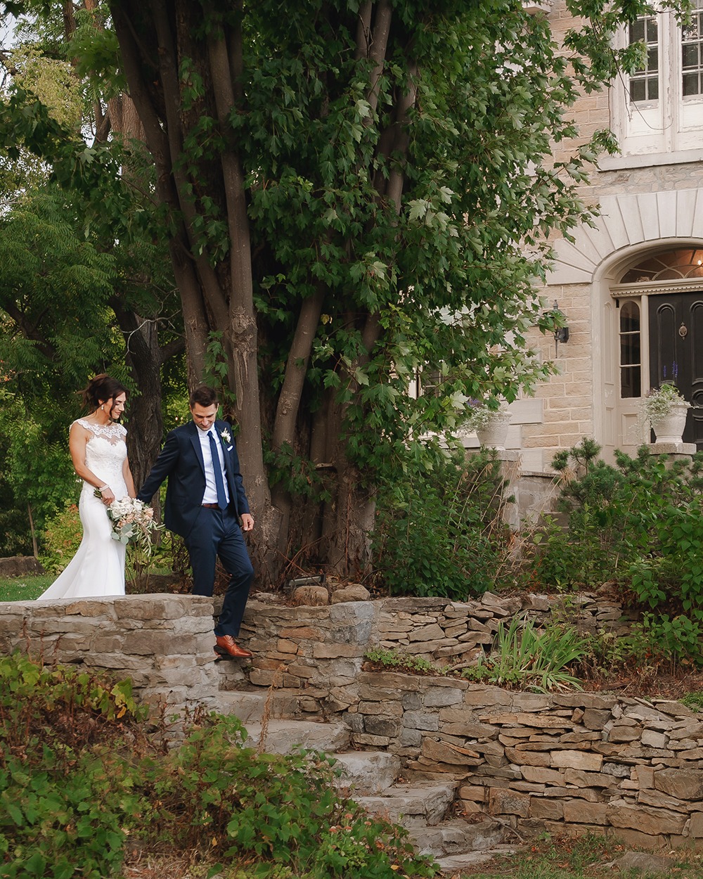 Bride and groom walking down steps together, with a large tree and farmhouse in the background