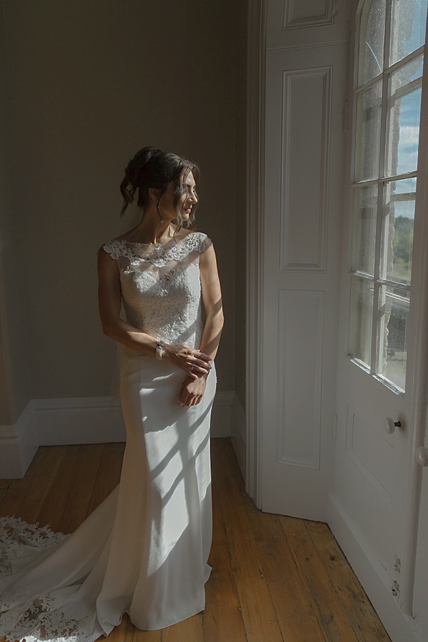 editorial bridal portrait. bride is holding her ams together facing camera while looking thorugh the window. soft lighting on her face