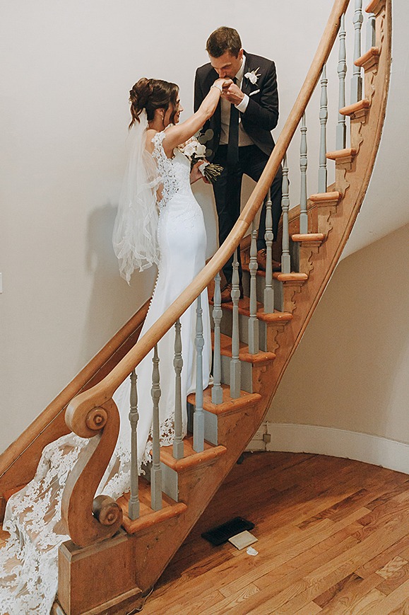 Bride and groom standing on vintage wooden farmhouse stairs, groom leaning down to kiss the bride’s hands.