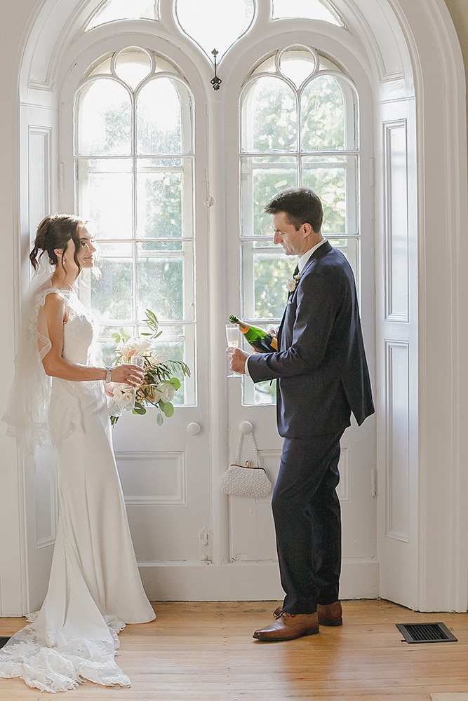 Bride and groom framed by a window at their farmhouse wedding, groom pouring champagne for a celebratory moment