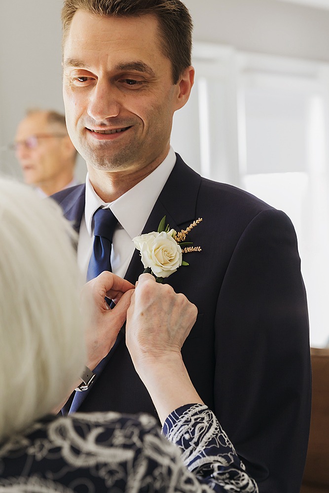 groom's mom is placing the buttenier onto groom's jacket while groom gently smiles at mom
